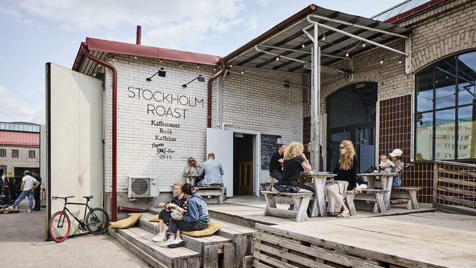 Folk sitter i en trappa och vid bord utanför en tegelbyggnad med texten "Stockholm Roast", foto. 