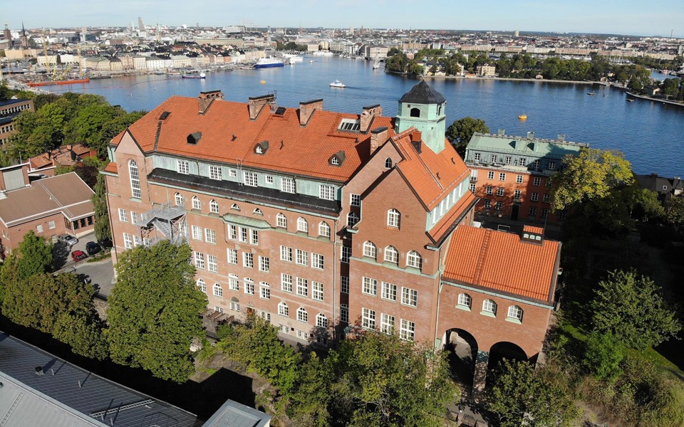 P A Fogelströms gymnasium ses snett uppifrån. Bakom skolar syns delar av Stockholm och Stockholms vatten, bland annat Gamla stan, Strandvägen, Djurgårdsfärjan och andra båtar och fartyg.