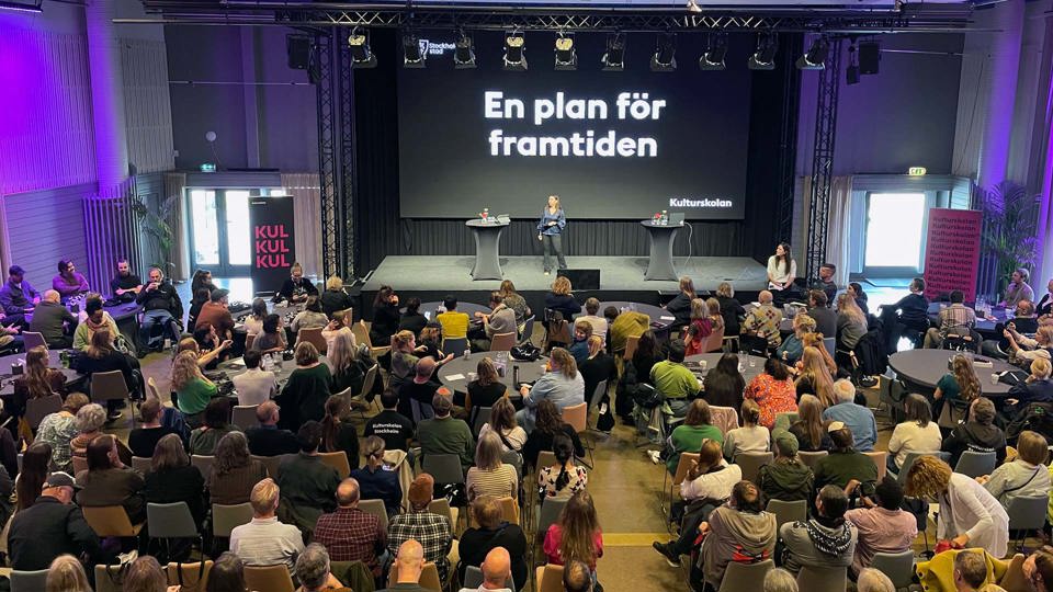 En stor sal med alla medarbetare samlade. På scenen står deras chef och pratar. 